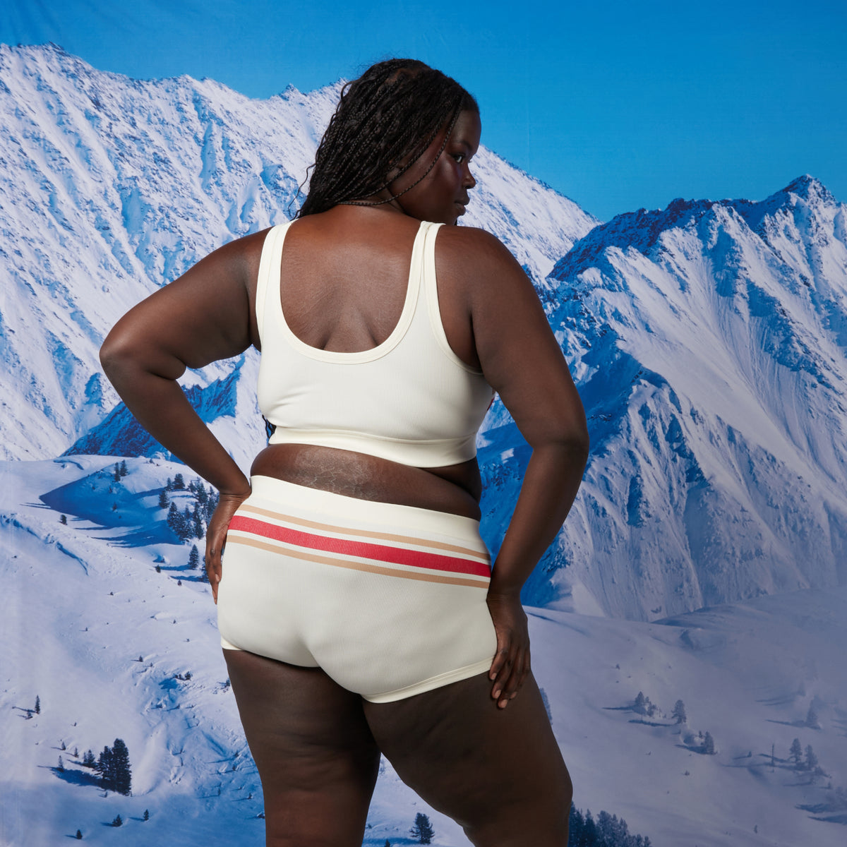 A woman stands with her back to the camera in front of a snowy mountain backdrop wearing a matching cream-colored bamboo rib underwear with red and tan stripes and small red jock tags. The set includes a cropped bra top and boy short style bottoms. Model: Rachel is 5'11" and wears a 2X
