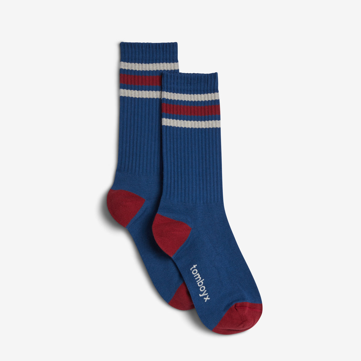 A pair of blue crew socks with red and white stripes near the cuff, red heels and toes, and the brand name 'tomboyx' printed in white on the sole of one sock.